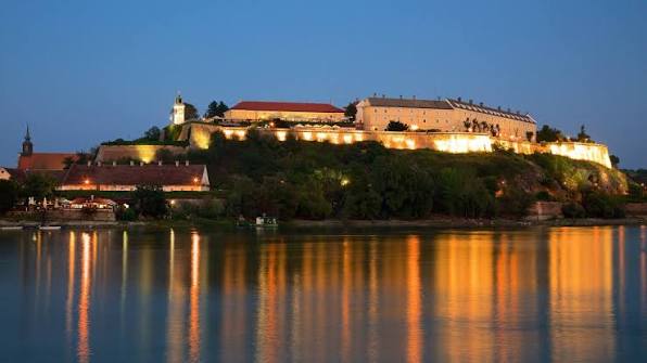Nighttime view of Petrovaradin Fortress in Novi Sad illuminated with golden lights reflecting on the Danube River.